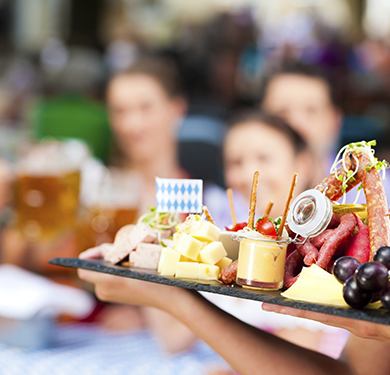 A waiter holding a flat cutting board packed with cheeses, meats, and fruit platter as a seated group of people wait in anticipation for it