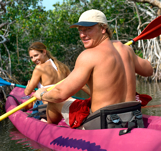 A couple sitting in a plastic two person kayak, paddling through mangroves, looking behind themselves at you