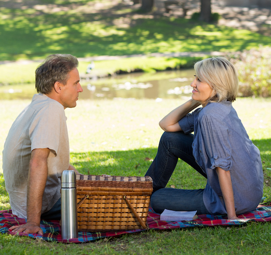 A couple sitting on a picnic blanket, with picnic basket and thermos next to them, gazing into each other's eyes