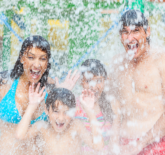 A family of four in bathing suits, smiling with their mouths open as they are getting soaked by the water falling on them in a water park